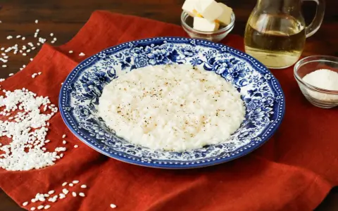 Un cremoso riso in bianco, condito con pepe nero, servito su un piatto decorato. Accanto, burro, olio e parmigiano grattugiato, ingredienti per un piatto semplice ma gustoso.