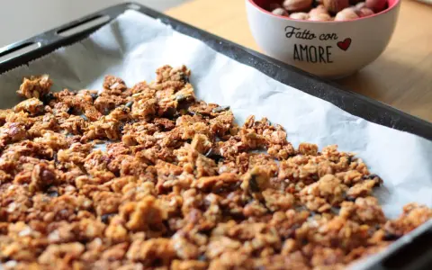 Granola fatta in casa appena sfornata su carta da forno, con una ciotola di frutta secca sullo sfondo.
