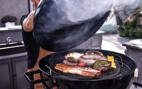 Un uomo solleva il coperchio di una grigliata di carne fumante, rivelando salsicce, pancetta, hamburger e verdure.
