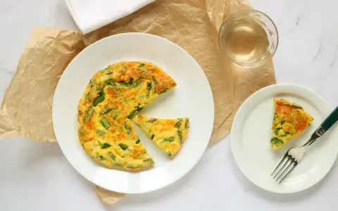 Deliziosa frittata di asparagi al forno, servita su un piatto bianco con una fetta a parte e un bicchiere di vino.