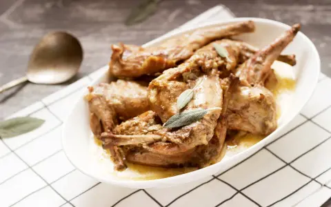 Un saporito coniglio in padella, cotto lentamente e guarnito con foglie di salvia, servito in un piatto bianco su una tovaglia a quadri.