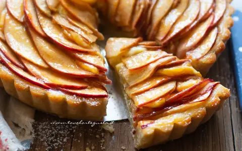 Una deliziosa crostata di mele, con fette di mela lucide disposte a spirale su una base dorata. Una fetta è stata tagliata, rivelando la morbidezza del ripieno.