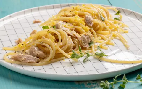 Un piatto di pasta tonno e limone, condita con scorza di limone grattugiata e erbe aromatiche fresche, servita su un piatto a scacchi bianchi.