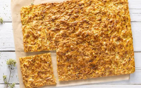 Schiacciata di zucchine dorata e croccante, tagliata a fette su carta da forno, con erbe aromatiche sparse.