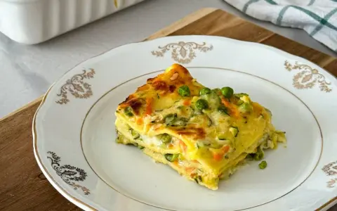 Una fetta di lasagne di verdure, con piselli e carote, su un piatto bianco decorato.