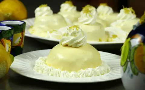Dolcetti rotondi con glassa al limone e panna montata, guarniti con scorza di limone. Sullo sfondo, limoni freschi e bicchierini decorati.