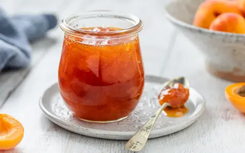 Vasetto di marmellata di albicocche, con un cucchiaino carico e albicocche fresche sullo sfondo.