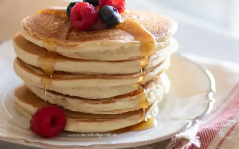 Pancakes senza uova dorati, impilati e inzuppati di sciroppo, guarniti con lamponi e mirtilli freschi.