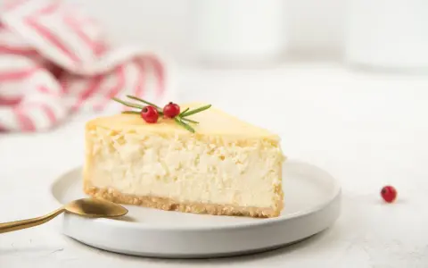 Una fetta di cheesecake yogurt greco, guarnita con bacche rosse e rosmarino, su un piatto bianco con un cucchiaio dorato.