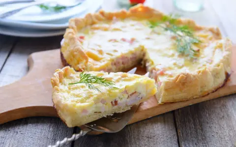 A slice of prosciutto and cheese tart is lifted from the whole tart on a wooden board, garnished with dill.