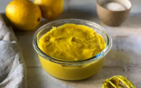 Crema pasticcera senza uova, densa e vellutata, in una ciotola di vetro. Limoni e zucchero sullo sfondo, pronti per un dolce delizioso.