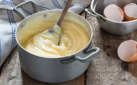 Crema pasticcera veloce in pentola, con cucchiaio di legno che mescola. Guscio d'uovo accanto, pronto per un dolce.