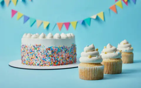Torta bianca con codette colorate e cupcake decorati con soffice crema al burro, su sfondo azzurro con festoni.