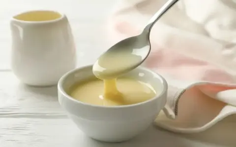 Un cucchiaio versa un denso latte condensato bimby in una ciotola bianca. Sullo sfondo, una piccola brocca e un tovagliolo rosa.