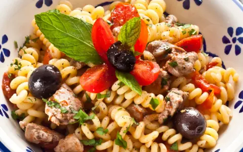 Pasta con pesce spada, pomodorini, olive nere e menta fresca in una ciotola decorata. Un piatto estivo che profuma di mare.