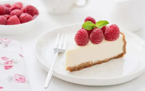 Una fetta di torta fredda allo yogurt guarnita con lamponi freschi e menta, su un piatto bianco con una forchetta.