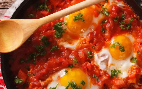 Uova al pomodoro cotte in padella, con tuorli dorati e prezzemolo fresco. Un cucchiaio di legno attende di servire questo piatto saporito.