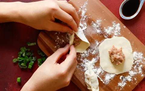 Mani che preparano il ripieno per ravioli su un tagliere di legno, con erba cipollina e salsa di soia a lato.