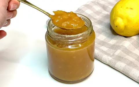 Un cucchiaio d'oro raccoglie la densa marmellata di limoni da un barattolo di vetro. Due limoni freschi riposano su un tovagliolo a quadretti.