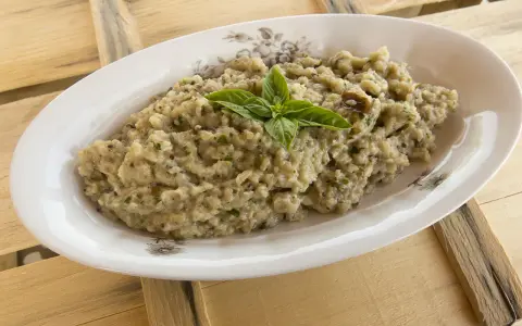 Crema di melanzane rustica, guarnita con basilico fresco, servita in un piatto ovale su un tavolo di legno grezzo.