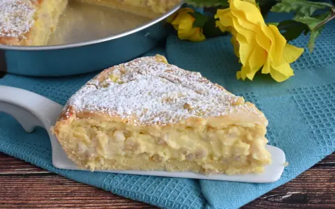 Una fetta di pastiera napoletana con crema pasticcera, cosparsa di zucchero a velo, su un piatto bianco, con fiori gialli sullo sfondo.