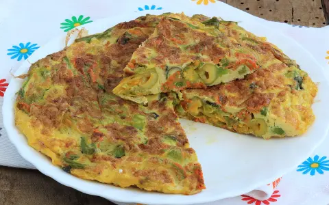 Frittata con fiori di zucca, carote e olive, tagliata a spicchi su un piatto bianco, con un tovagliolo fiorato sullo sfondo.
