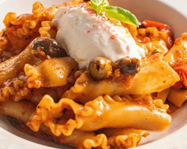 Pasta con melanzane, condita con un sugo ricco, olive e una generosa quenelle di ricotta fresca, guarnita con basilico.