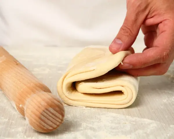 Mani che piegano un impasto per pasta fatta in casa, con un mattarello di legno accanto, cosparso di farina.