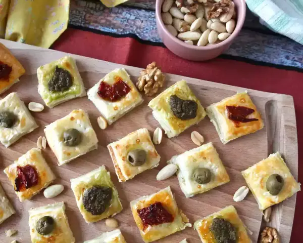 Sfiziose ricette con pasta sfoglia: quadratini dorati con olive, pesto e pomodori secchi, perfetti per un aperitivo.