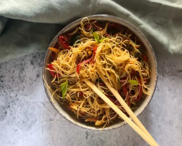 Un piatto invitante di spaghetti di riso con verdure colorate, come peperoni rossi e zucchine, guarnito con basilico fresco e servito con bacchette.