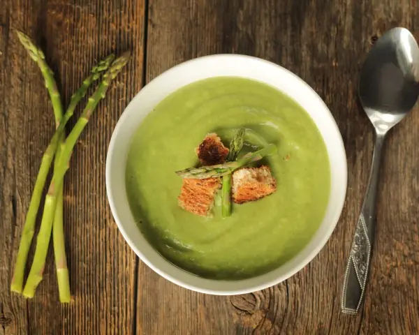 Una ciotola di cremosa crema di asparagi, guarnita con crostini e germogli freschi, accanto a un mazzo di asparagi su un tavolo di legno.