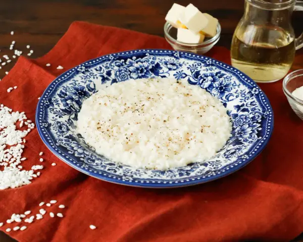 Un cremoso riso in bianco, condito con pepe nero, servito su un piatto decorato. Accanto, burro, olio e parmigiano grattugiato, ingredienti per un piatto semplice ma gustoso.