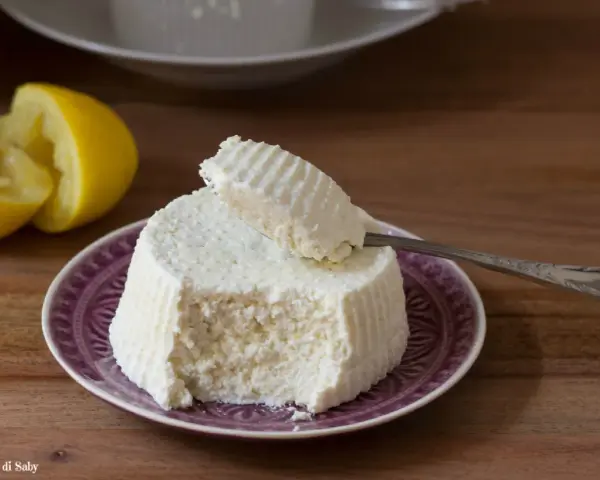 Ricotta fatta in casa su un piatto viola, con limone tagliato a metà sullo sfondo. Un cucchiaino preleva una porzione cremosa.