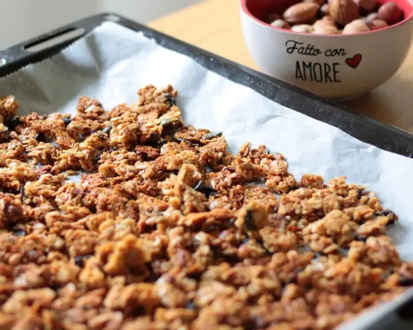 Granola fatta in casa appena sfornata su carta da forno, con una ciotola di frutta secca sullo sfondo.