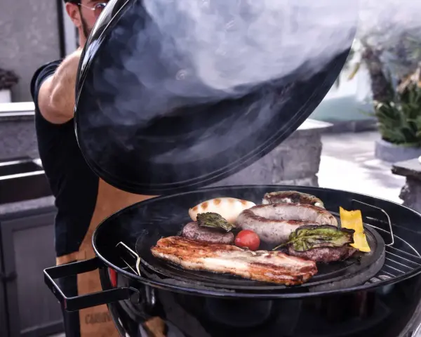 Un uomo solleva il coperchio di una grigliata di carne fumante, rivelando salsicce, pancetta, hamburger e verdure.