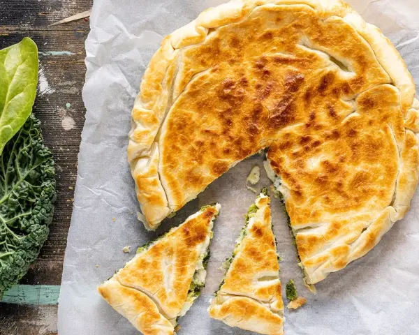 Torta salata con pasta sfoglia dorata, ripiena di verdure. Accanto, foglie di cavolo nero fresco.
