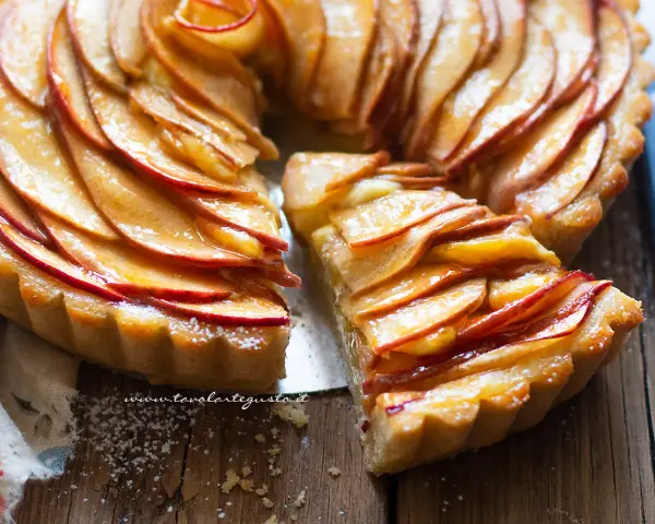 Una deliziosa crostata di mele, con fette di mela lucide disposte a spirale su una base dorata. Una fetta è stata tagliata, rivelando la morbidezza del ripieno.