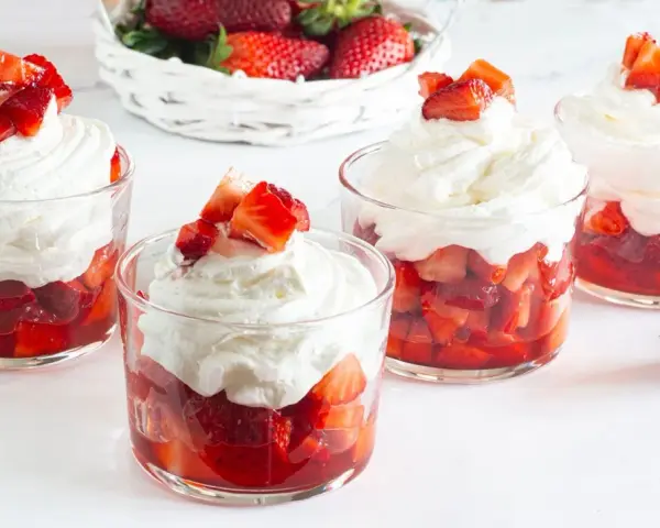 Coppette di fragole con panna montata e pezzetti di fragola fresca in cima. Un cestino di fragole sullo sfondo.