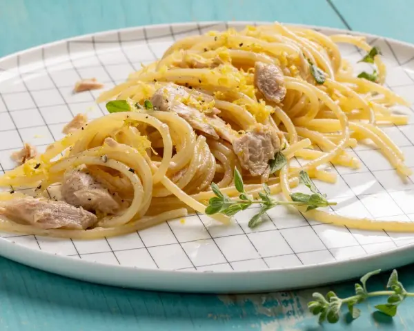 Un piatto di pasta tonno e limone, condita con scorza di limone grattugiata e erbe aromatiche fresche, servita su un piatto a scacchi bianchi.