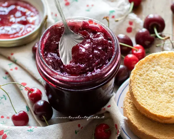 Un barattolo pieno di deliziosa marmellata di ciliegie, con ciliegie fresche e fette biscottate accanto.