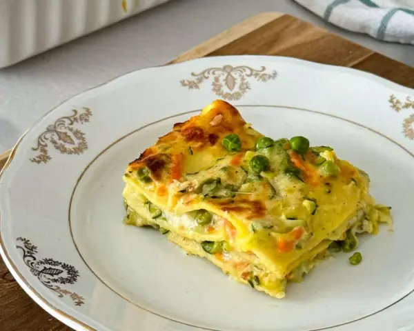 Una fetta di lasagne di verdure, con piselli e carote, su un piatto bianco decorato.
