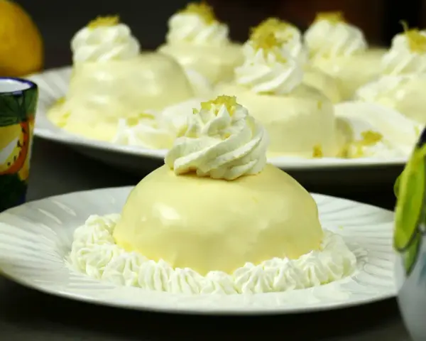 Dolcetti rotondi con glassa al limone e panna montata, guarniti con scorza di limone. Sullo sfondo, limoni freschi e bicchierini decorati.
