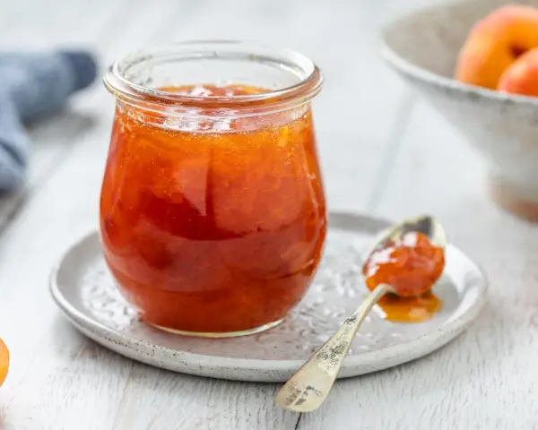 Vasetto di marmellata di albicocche, con un cucchiaino carico e albicocche fresche sullo sfondo.