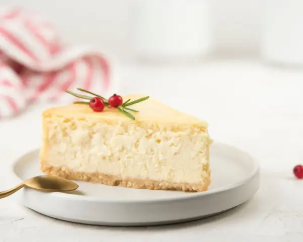 Una fetta di cheesecake yogurt greco, guarnita con bacche rosse e rosmarino, su un piatto bianco con un cucchiaio dorato.