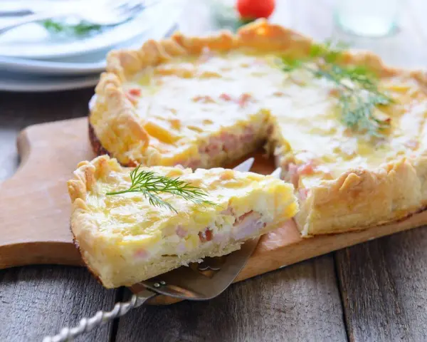A slice of prosciutto and cheese tart is lifted from the whole tart on a wooden board, garnished with dill.