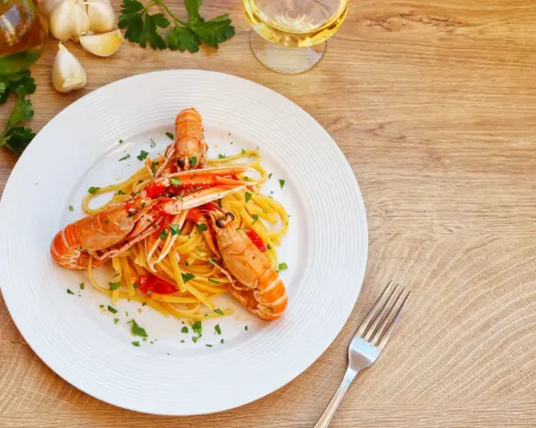 Un piatto di linguine agli scampi, condite con pomodorini e prezzemolo, accompagnato da ingredienti freschi e un bicchiere di vino bianco.