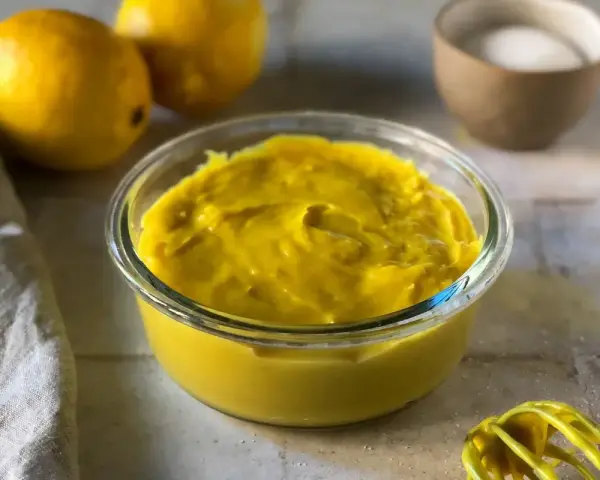 Crema pasticcera senza uova, densa e vellutata, in una ciotola di vetro. Limoni e zucchero sullo sfondo, pronti per un dolce delizioso.