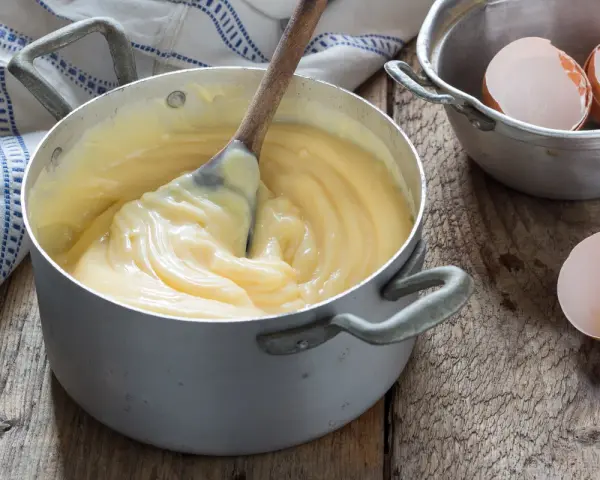 Crema pasticcera veloce in pentola, con cucchiaio di legno che mescola. Guscio d'uovo accanto, pronto per un dolce.