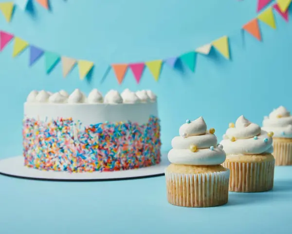 Torta bianca con codette colorate e cupcake decorati con soffice crema al burro, su sfondo azzurro con festoni.