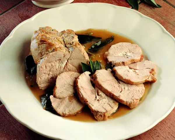 Delizioso arrosto di tacchino affettato, servito in un sugo saporito con foglie di salvia su un piatto bianco.